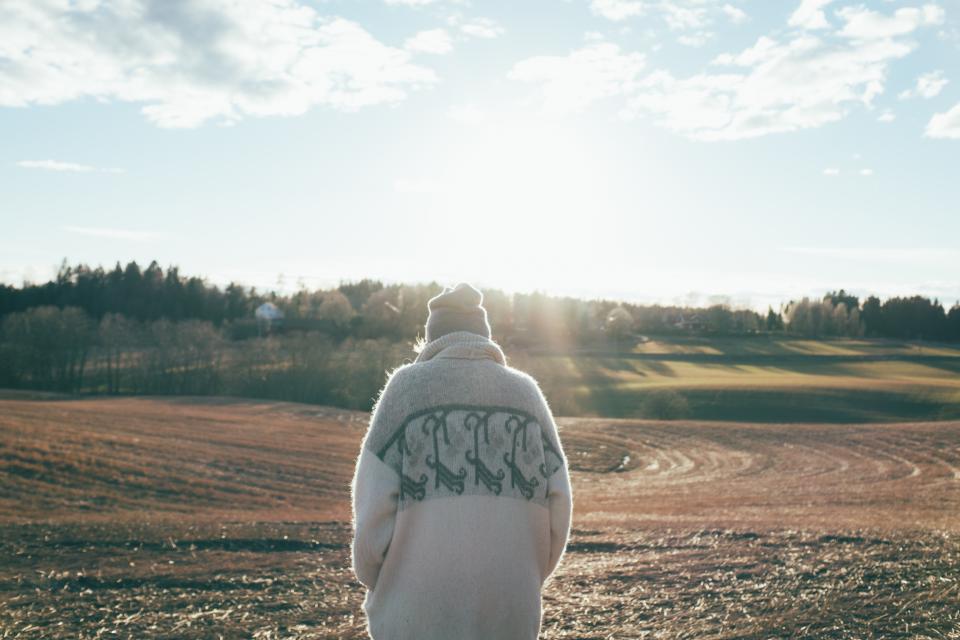 Person with their back facing the camera, wearing a beige wool sweater and a grey hat. The person is standing in a field on a sunny day. 